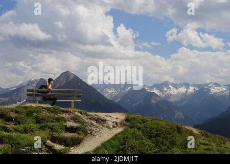Eine junge Frau macht eine kurze Pause auf einer Bank hoch über den Bergen Stockfoto