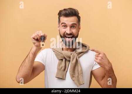 Aufgeregt Mann hält Schlüssel und jubelte, zeigt mit dem Finger auf sie, bärtige Kerl jubelte isoliert auf orangenen Hintergrund, zog in neue Wohnung Stockfoto