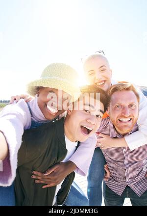 Vertikales Bild Piggyback-Freunde nehmen an einem sonnigen Tag zusammen ein lächelndes Selfie im Freien auf. Portrait Gruppe von jungen Menschen, die Spaß zusammen haben. Jungs Stockfoto