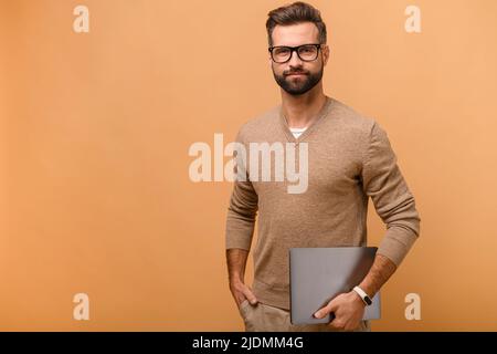 Porträt eines ernsthaften, intelligenten bärtigen Mannes in einer Brille, der Laptop-Computer isoliert auf beigem Hintergrund hält, stolzer männlicher Unternehmer, der die Kamera anschaut Stockfoto