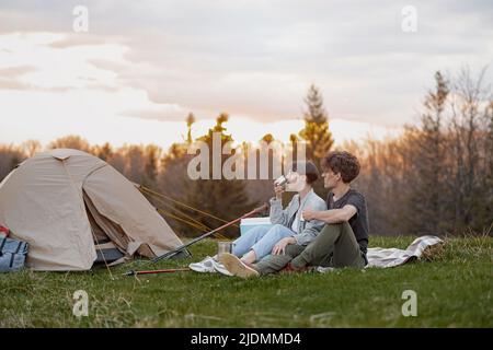 Junges Paar, das sich in der Natur ausruhte, mit Zelt, sitzend, sprechend und Tee oder Kaffee trinkend. Stockfoto