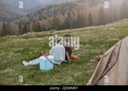 Hinter einem romantischen Paar, das auf Gras im Zelt hoch in den Bergen sitzt. Wunderschöne Aussicht. Stockfoto