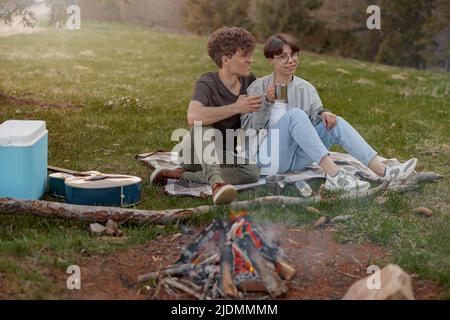 Fröhliches Paar sitzt auf Gras beim Picknick, redet und trinkt Tee oder Kaffee. Tourismus. Stockfoto