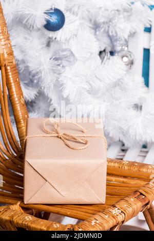Das Geschenk ist in Kraftpapier und Kordelzug auf einem Korbsessel auf einem Hintergrund von weiß dekorierten künstlichen Weihnachtsbaum gewickelt. Stockfoto