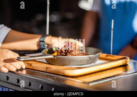 Kellner hält Holztablett mit frisch gekochtem Fleisch in einer Pfanne auf einem Holztisch im Luxusrestaurant. Gegrillter Brot mit Gemüse und Koch auf Stockfoto