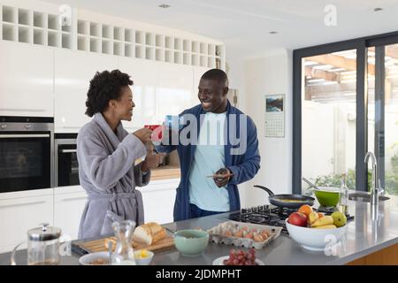 Fröhliches afroamerikanisches Paar mit mittleren Erwachsenen, das in der Küche Bademäntel und eine Tasse Kaffee trägt Stockfoto