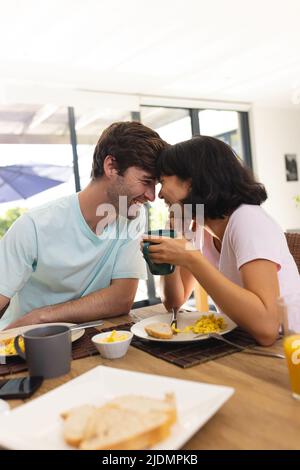 Glückliches, multirassisches junges Paar mit Kopf-an-Kopf-Romanze beim Frühstück am Esstisch Stockfoto