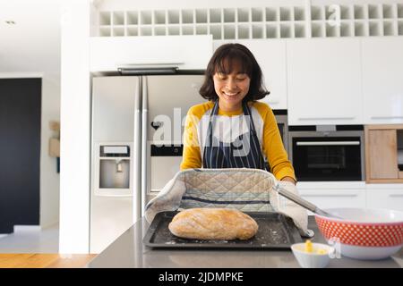 Lächelnde asiatische junge Frau mit kurzen Haaren hält frisch gebackenes Brot auf Kücheninsel Stockfoto