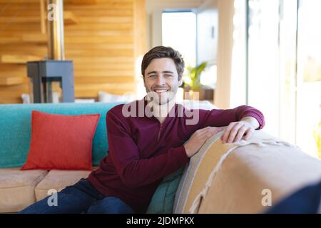 Porträt eines lächelnden, hübschen kaukasischen jungen Mannes, der im Wohnzimmer auf dem Sofa sitzt Stockfoto