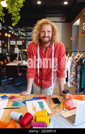 Porträt eines lächelnden kaukasischen männlichen Modedesigners, der sich am Schreibtisch im Studio lehnt Stockfoto