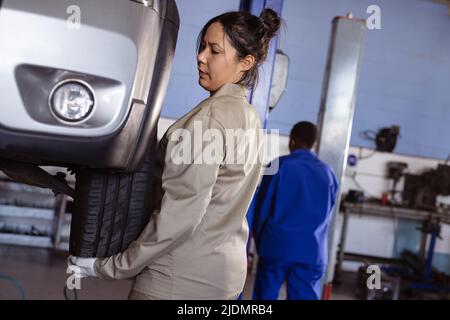 Mittlere Erwachsene asiatische Mechanikerin, die Autoreifen in der Werkstatt mit einer Kollegin im Hintergrund repariert Stockfoto