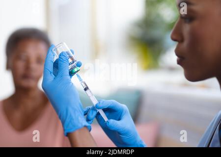 afroamerikanische ältere Frau, die eine junge Ärztin ansieht, die zu Hause Fläschchen und Spritze hält Stockfoto
