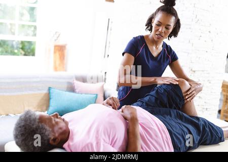 afroamerikanische Physiotherapeutin massiert das Bein einer älteren Frau, die zu Hause auf dem Tisch liegt Stockfoto