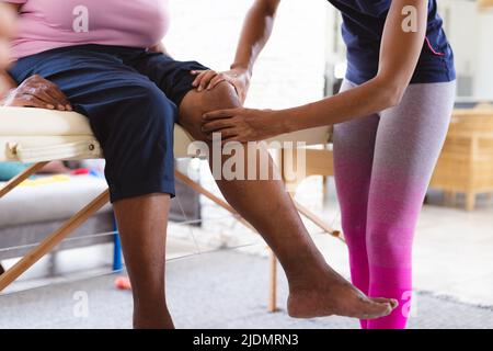 afroamerikanische Physiotherapeutin massiert zu Hause das Knie einer älteren Frau Stockfoto