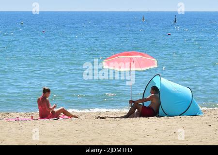 Paar am Strand mit rotem Sonnenschirm und blauem Strandzelt, Boscombe Beach, Bournemouth, Dorset, UK, Juni 2022, heißes Wetter am Nachmittag in der Sommersonne. Stockfoto