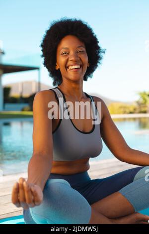 Fröhliche junge afroamerikanische Afro-Frau, die Yoga macht, während sie am Pool sitzt und Platz kopiert Stockfoto