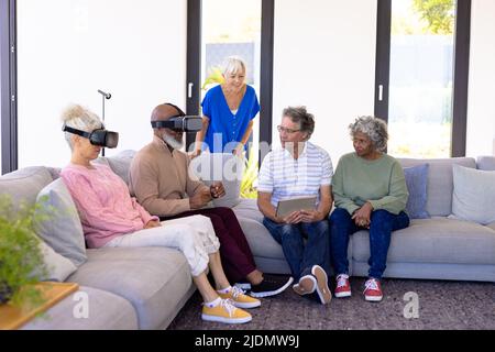 Multirassische Senioren mit digitalem pc, die Freunde mithilfe des Virtual-Reality-Simulators auf dem Sofa betrachten Stockfoto