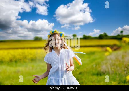 Schönes, energiegeladenes, verträumtes Teenager-Mädchen in einem weißen Kleid und einem leuchtend ukrainischen Kranz, das durch gelb blühende Rapsfelder und grüne Äcker läuft Stockfoto