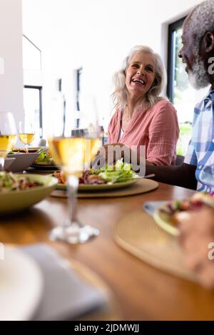 Fröhliches, multirassisches Seniorenpaar, das sich beim Mittagessen am Esstisch im Pflegeheim die Hände hielt Stockfoto