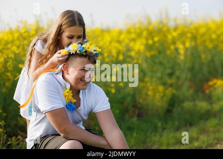 Eine fröhliche jüngere Schwester flechzt lange, dünne, mehrfarbige Bänder in einem ukrainischen gelb-blauen Kranz mit hellen wilden Blumen auf dem Kopf des älteren Bruders, Stockfoto