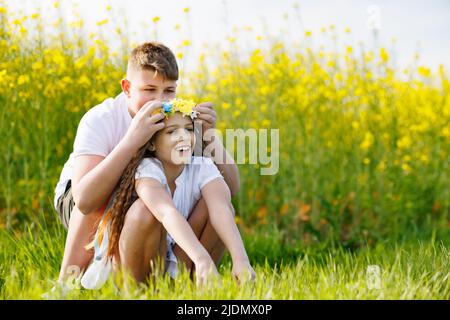 Der Bruder des Teenagers flechzt mehrfarbige Bänder in einem gelb-blauen Kranz mit wilden Blumen auf dem Kopf der fröhlich lächelnden Schwester auf der grünen Wiese A Stockfoto