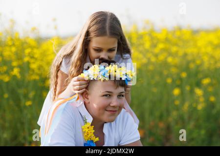 Eine fröhliche jüngere Schwester flechzt lange, dünne, mehrfarbige Bänder in einem ukrainischen gelb-blauen Kranz mit hellen wilden Blumen auf dem Kopf des älteren Bruders, Stockfoto