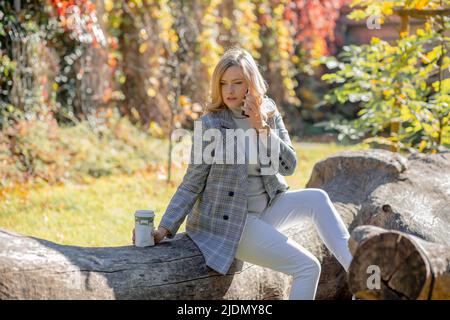Schöne Blondine in leichtem Pullover und Tweed-Jacke spricht am Telefon sitzen auf einem gefallenen Baum an sonnigen Herbsttag im Park. Herbststimmung. Kaffeepause Stockfoto