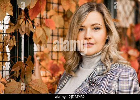 Gepflegte, charmante, 35-40 Jahre alte Blondine mit langen Haaren in leichtem Pullover und Tweed-Jacke an der Wand aus roten Herbstblättern im Stadtgarten Stockfoto