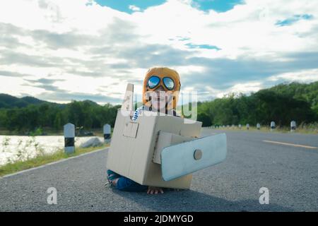 Nettes Träumer kleines Mädchen, das an einem sonnigen Tag mit Pappflugzeugen auf einer Seestraße spielt. Glückliches Kind spielt mit Pappflugzeug gegen blauen Sommerhimmel b Stockfoto