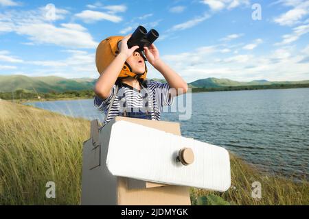 Niedliches Träumerin kleines Mädchen, das an einem sonnigen Tag mit Pappflugzeugen und Ferngläsern auf einer Wiese am See spielt. Kindheitstraum Imagination Konzept. Stockfoto