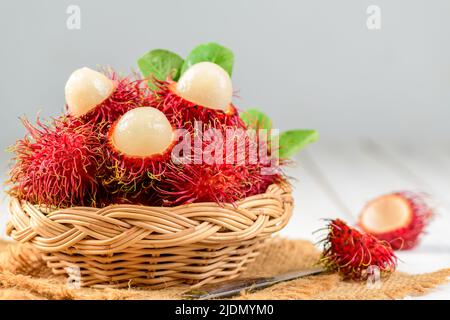 Frische Rambutan-Früchte mit Blättern im Bambuskorb auf Holzhintergrund. Obst Südostasien in der Sommersaison Stockfoto