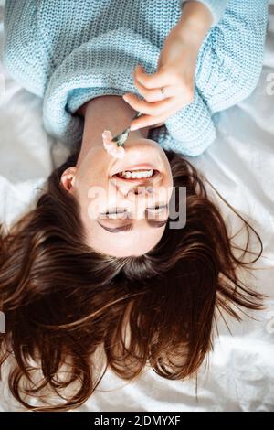 Nette junge Frau in blauem Pullover mit langen dunklen Haaren, die auf dem Bett mit Blume liegt und Spaß hat Stockfoto