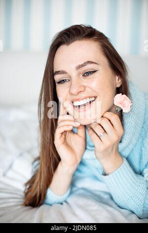 Romantische junge Frau mit toothy Lächeln in blauen Pullover liegend auf dem Bett mit Blume in der Hand. Porträt einer verträumten Dame, die sich ausruhte Stockfoto