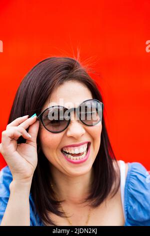 Junge Brünette Frau spielt mit ihrer Sonnenbrille vor einem roten Hintergrund Stockfoto