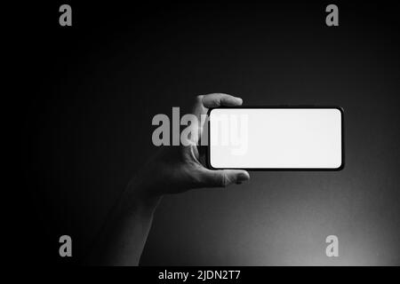 Schwarz-weißes Bild der Hand des Mannes hält schwarzes Smartphone horizontal mit leeren weißen Bildschirm isoliert auf dunklem Hintergrund mit dramatischer Beleuchtung ein Stockfoto