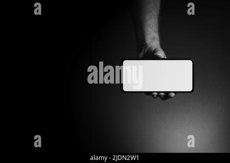 Schwarz-weißes Bild der Hand des Mannes hält schwarzes Smartphone horizontal mit leeren weißen Bildschirm isoliert auf dunklem Hintergrund mit dramatischer Beleuchtung ein Stockfoto