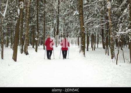 Frauen mit Wanderstöcken im Winterwald. Rennen zu Fuß im Park im Winter. Rentner spielen Sport. Großmütter in roten Mänteln. Frauen verlassen sich auf Skistöcke. Stockfoto