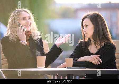 Traurige Frau, die von ihrer Freundin ignoriert wird, die im Straßencafé im Freien sitzt, während sie glücklich auf dem Handy redet und keine Aufmerksamkeit schenkt. Freundschaft Stockfoto