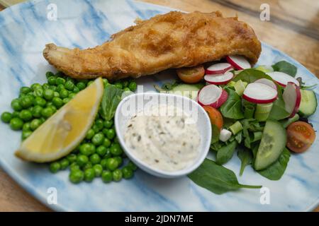 Teig auf einem Kabeljaufisch auf einem Teller. Im flachen Fokus ist ein Salat und Erbsen. Es gibt auch eine Zitronenscheibe und einen Topf mit Tartarsauce, alles auf einem blauen und weißen pla Stockfoto