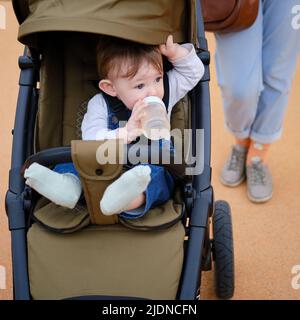 Frau Mutter geht mit Kleinkind Baby Junge sitzt in einem Kinderwagen auf dem Spielplatz des Parks. Kind in einem Kinderwagen mit Mama. Kind im Alter von einem Jahr Stockfoto