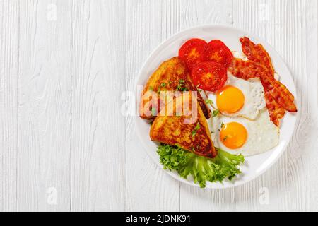 irisches Frühstück mit Kartoffelpüree, Spiegeleiern, Speckscheiben, gegrillten Tomaten und frischem Salat auf weißem Teller auf weißem Holztisch, horizontal Stockfoto