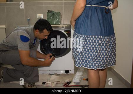 Bild eines Gerätetechnikers, der eine defekte Waschmaschine repariert und einer Frau das Problem erklärt. Stockfoto