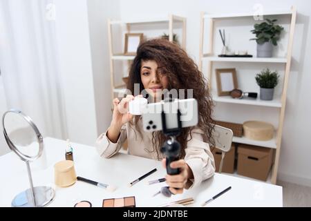 Selbstbewusst gebräunt gut aussehende lockige lateinische Schönheit Blogger in Leinenhemd wirbt effektive Creme in Hause weiß Interieur. Space Mockup Banner kopieren Stockfoto