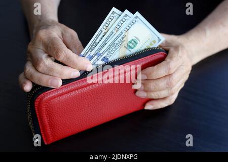 Ältere Frau nimmt US-Dollar aus der roten Brieftasche, faltige weibliche Hände aus der Nähe. Konzept der Pensionszahlungen, Ersparnisse, Rentner in den USA Stockfoto