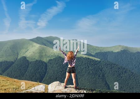 Das junge Mädchen steht mit einem Rucksack auf einem Berg, spreizt die Arme zur Seite und genießt den Blick auf die Berge, die mit Grün und weißem Kl. bedeckt sind Stockfoto