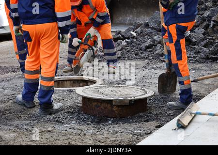 Arbeiter reparieren den Kanal, der sich gut an der Stadtstraße befindet, männliche Beine in der Nähe des Schachtkanals. Bau- und Installationsarbeiten Stockfoto