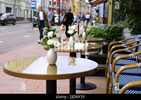 Straßencafé in der Sommerstadt mit leeren, nassen Tischen im Freien im Hintergrund von Wanderern. Vasen aus Rosenblumen auf runden Tischen und Stühlen nach Regen Stockfoto