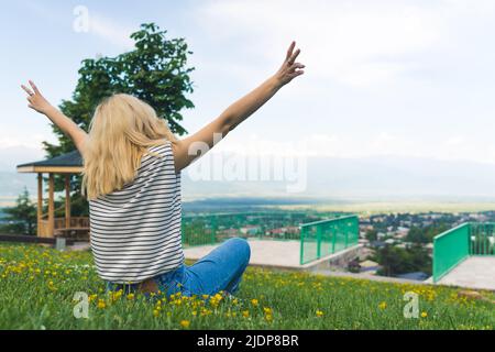 Mittlere Aufnahme eines blonden Mädchens, das auf dem grünen Gras sitzt und eine Aussicht genießt. Hochwertige Fotos Stockfoto