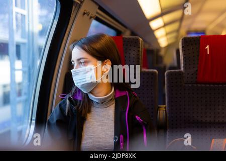 Frau in Maske sitzend im Zug und mit Blick aus dem Fenster Stockfoto
