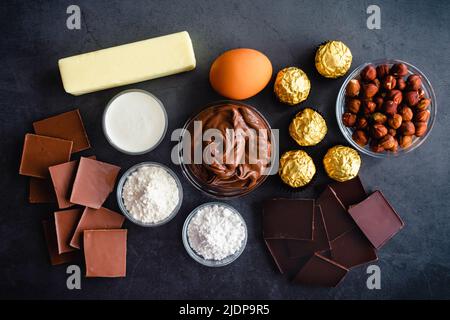 Schokoladen-Haselnuss-Torte Zutaten von oben gesehen: Schokolade, Haselnüsse, Butter und andere Dessertzutaten auf dunklem Hintergrund Stockfoto